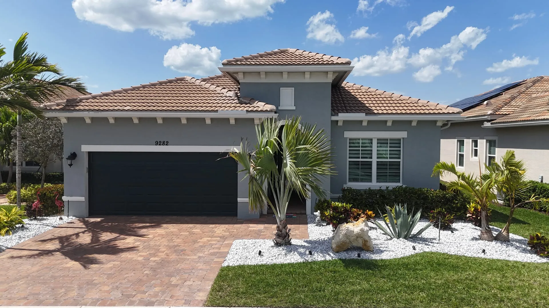 Drone photo of a gray stucco home with a tile roof, palm trees and white rock landscaping