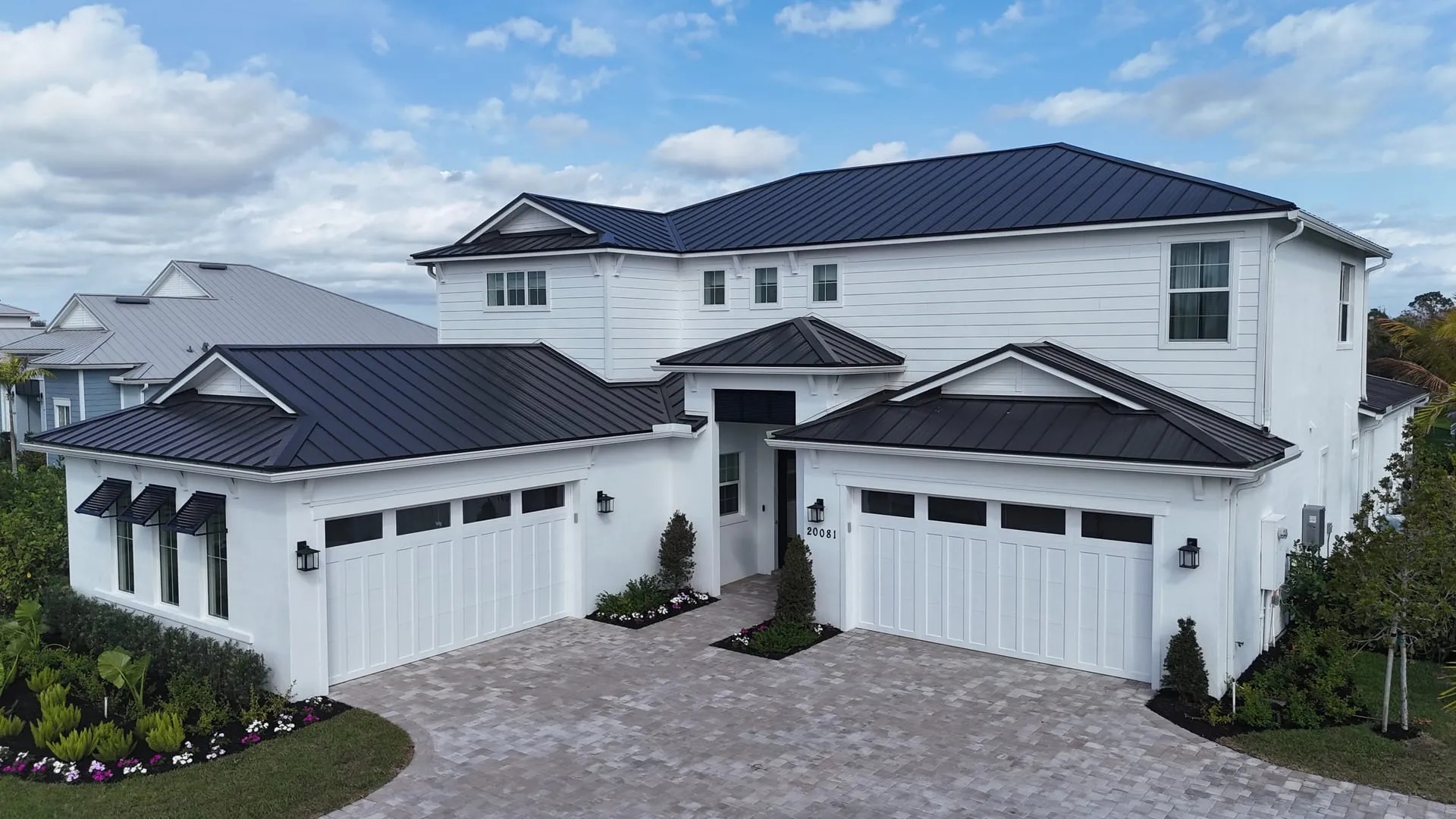 Drone photo of a white modern farmhouse-style home with a dark metal roof, two garages and a paver driveway under a blue sky