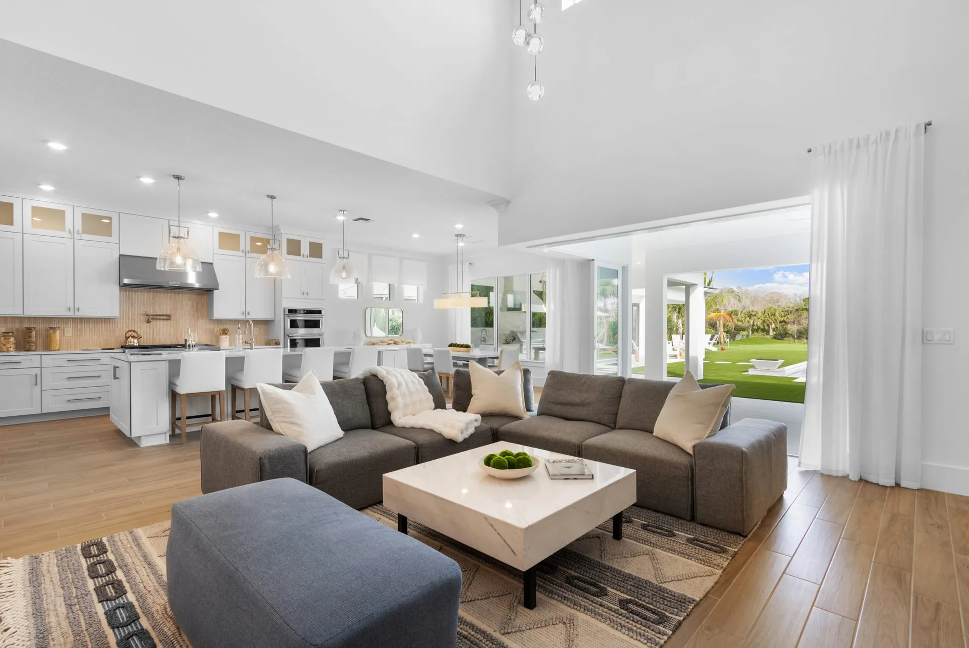 Open great room with a gray sectional sofa, a white kitchen behind it and sliding doors to the lawn