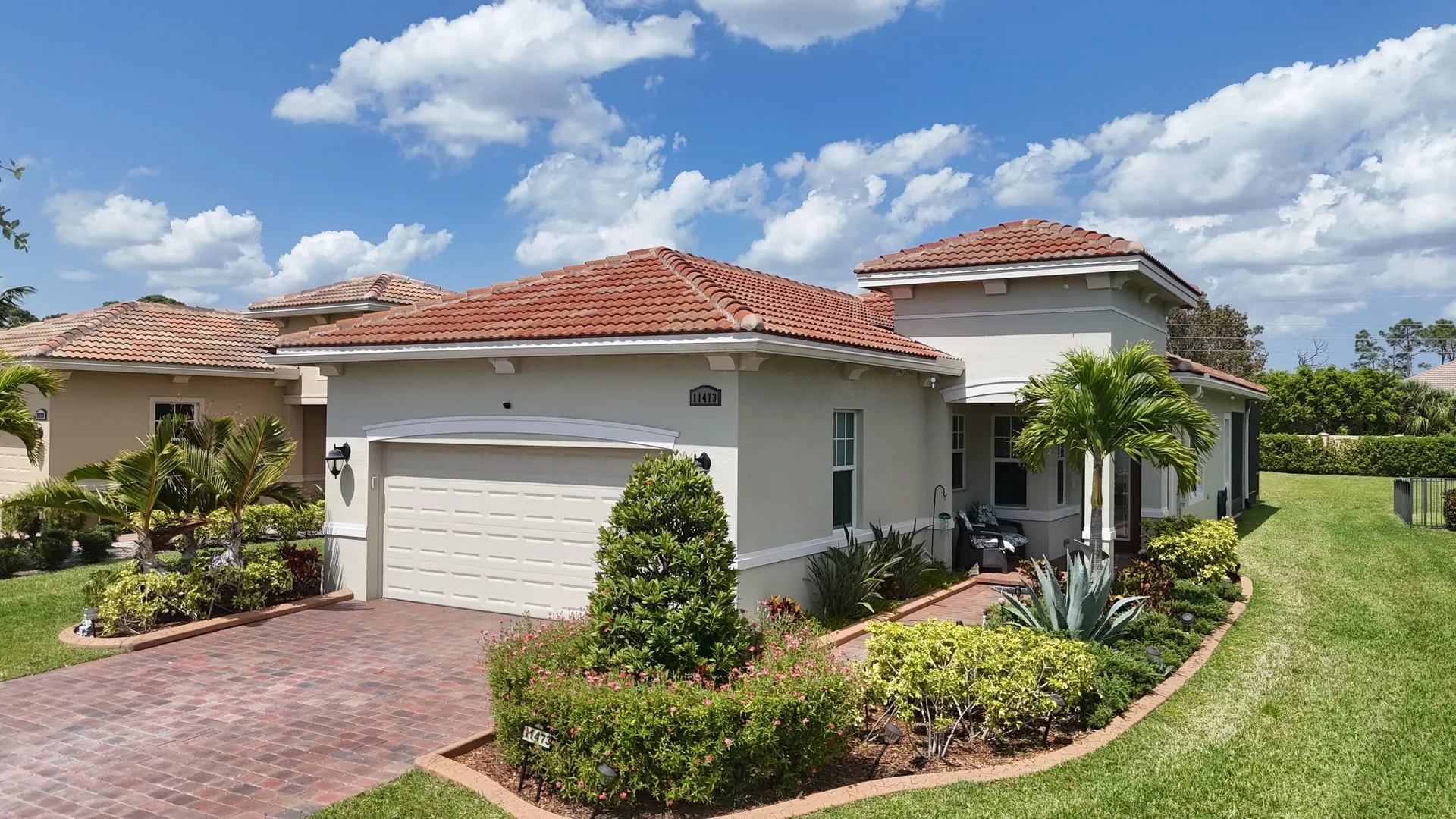Drone photo of a tan stucco home with a terracotta tile roof, palm trees and a red paver driveway