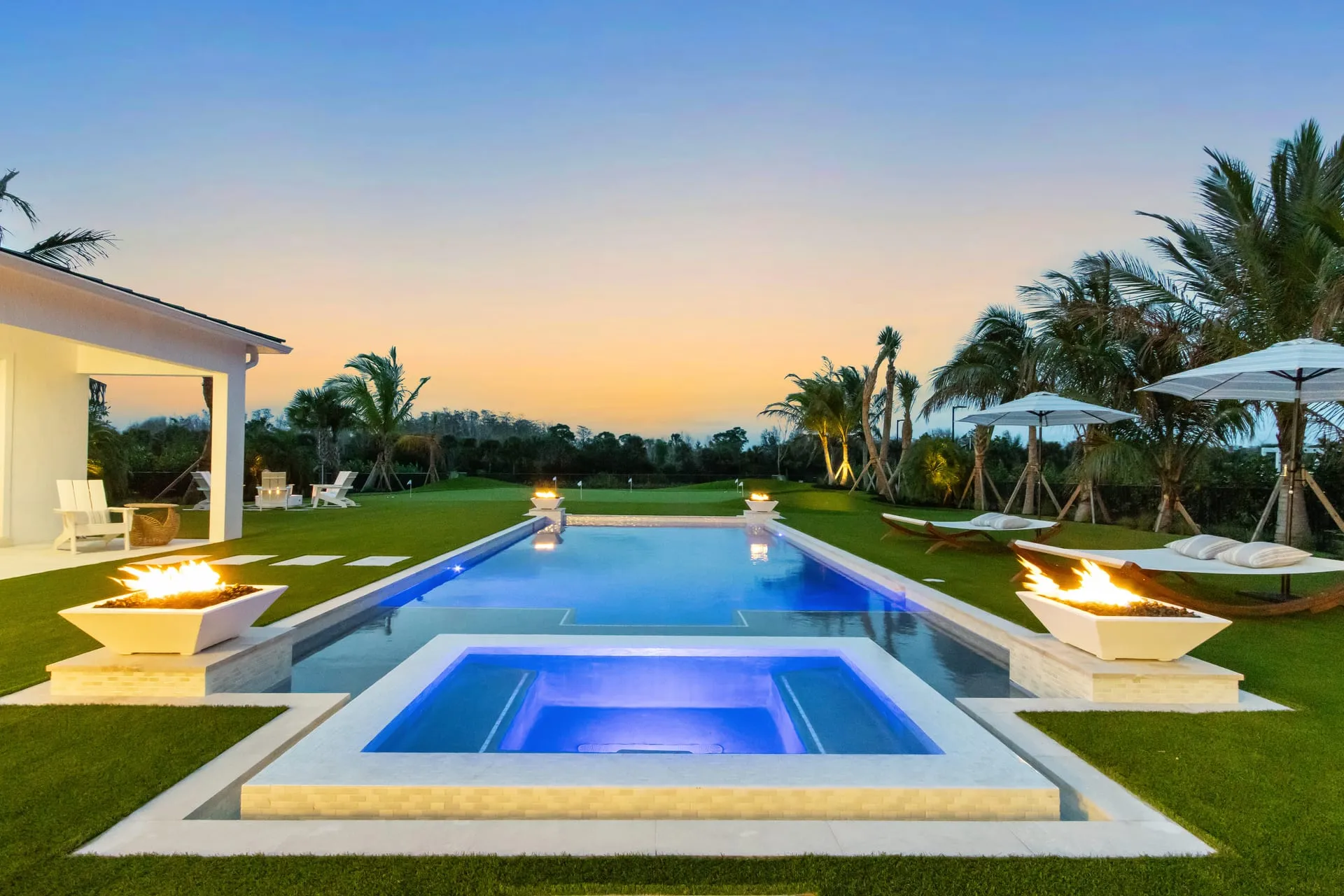 Backyard pool and spa at twilight with fire bowls, palm trees and patio umbrellas under a sunset sky