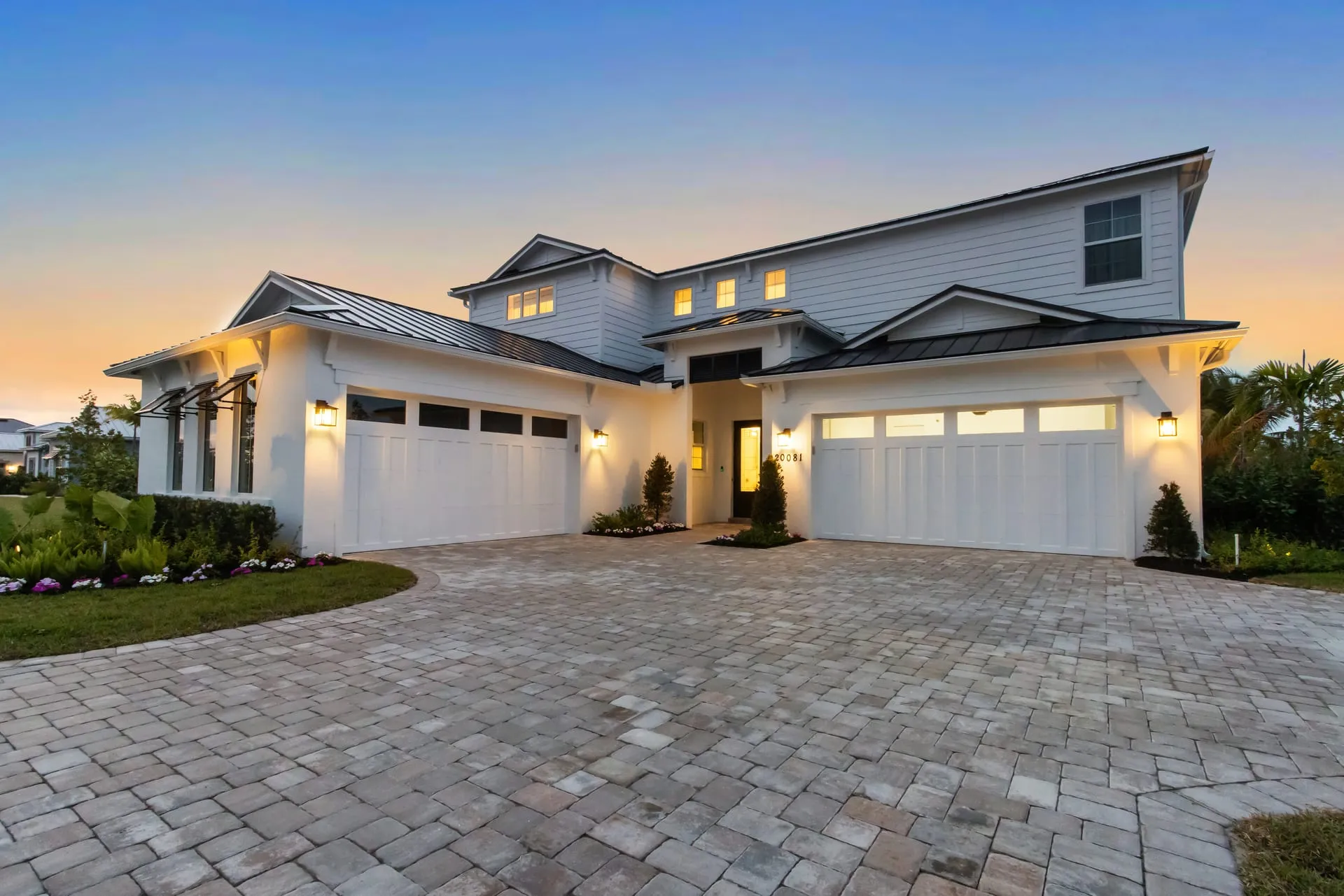 White modern farmhouse-style home at twilight with glowing windows, lit garage doors, a paver driveway and a sunset sky